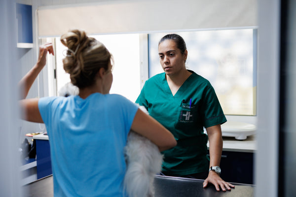 Two healthcare professionals in a clinical setting, one in a blue shirt and the other in green scrubs.